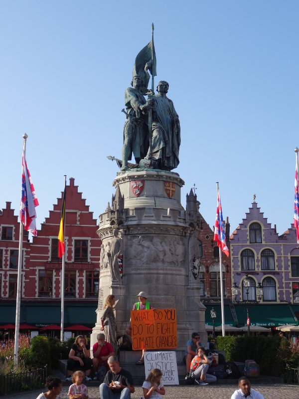 Statue der historischen Aufrührer Breydel und de Coninck auf dem Marktplatz von Brügge, im Hintergrund eine Häuserzeile, unter der Statue Menschen, die für den Klimaschutz demonstrieren.